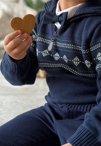 Sini neule, jossa on geometrisia kuvioita ja vetskari, pitää sydämen muotoista piparkakkua. Laita nuoli ja tummat housut täydentävät ilmeen.