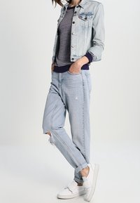 Veste en denim bleu clair avec fermeture à boutons, pull en tricot blanc, jean bleu clair déchiré, et baskets blanches avec des accents en nœud.