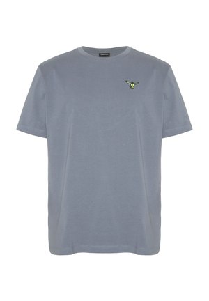 Mann mit orangefarbener Kappe, grauem T-Shirt mit kleinem grünem Logo, schwarzen Shorts mit Kordelzug und weißen Turnschuhen, steht und schaut zur Seite.