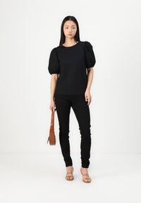 Svart puffermed topp med svarte skinny jeans. Modellen holder en liten brun håndveske. Studioinnstilling med en nøytral bakgrunn.
