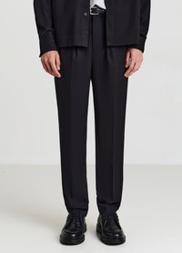 Schwarze, maßgeschneiderte Hose mit geradem Bein, aus glattem Stoff gefertigt. Kombiniert mit schwarzen Schnürschuhen und einem dunklen Hemd mit Knöpfen.