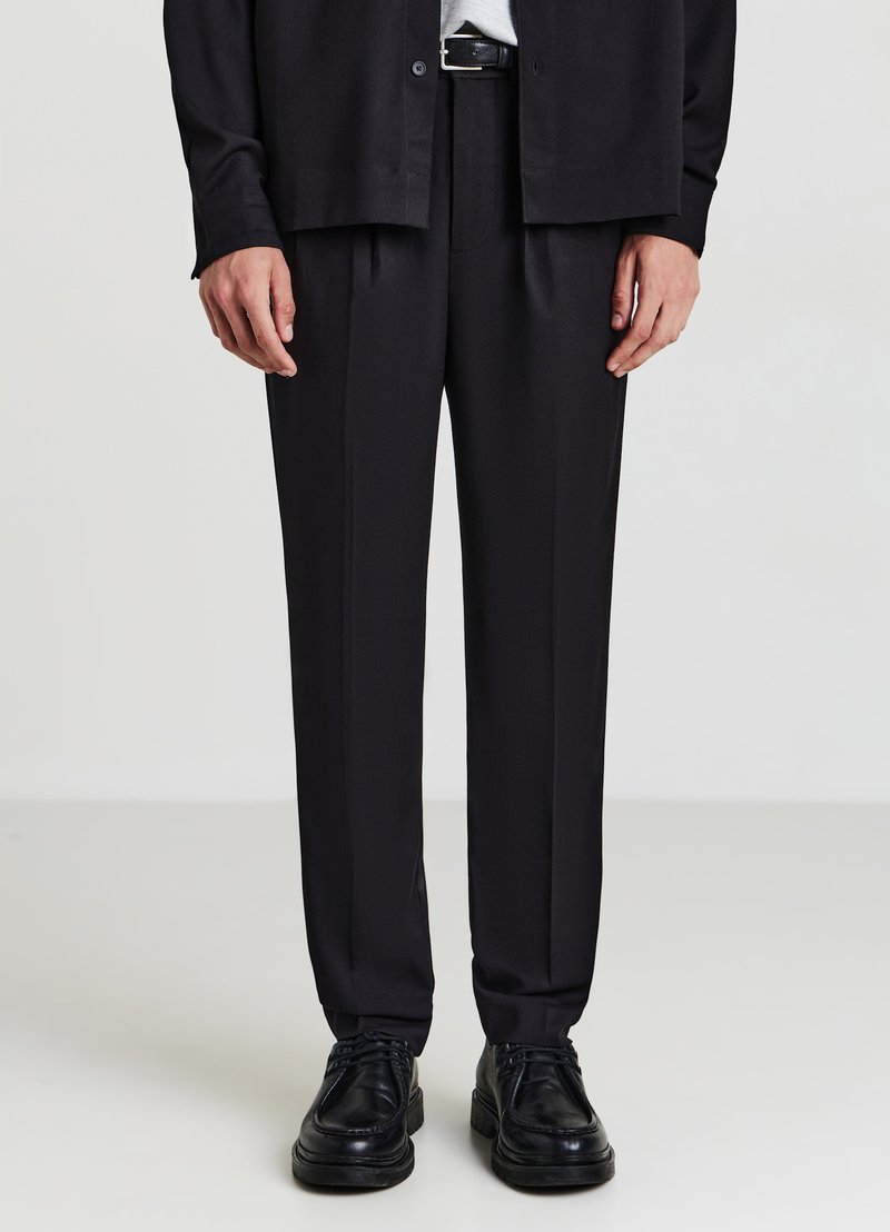 Schwarze, maßgeschneiderte Hose mit geradem Bein, aus glattem Stoff gefertigt. Kombiniert mit schwarzen Schnürschuhen und einem dunklen Hemd mit Knöpfen.