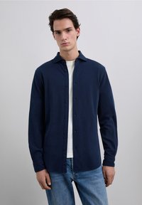 Chemise à manches longues bleu marine avec une texture douce, col à pointes, et un logo discret sur la poitrine gauche, portée avec un jean en denim clair.