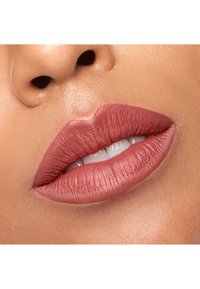Rouge à lèvres mat dans une teinte rose chaud, texture lisse, appliqué sur des lèvres pleines, avec un éclat naturel sur les dents et la peau. Vue en gros plan.