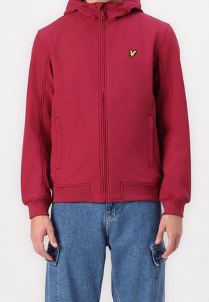 Mężczyzna ubrany w bordową kurtkę zapinaną na zamek z kapturem i żółtym logo ptaka na piersi, w parze z niebieskimi jeansowymi spodniami typu cargo.