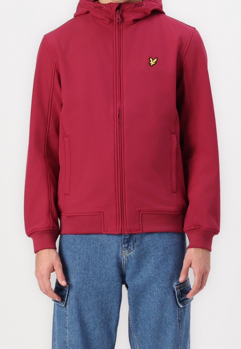 Homme portant une veste bordeaux zippée avec capuche et logo d'oiseau jaune sur la poitrine, associée à un pantalon cargo en denim bleu.