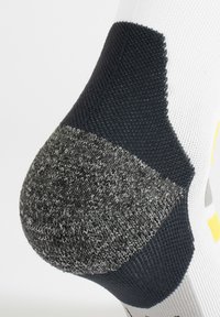 Calza bianca con tallone grigio rinforzato e accenti blu navy; texture a strisce nella zona del tallone, con motivi gialli e grigi brillanti sul corpo.