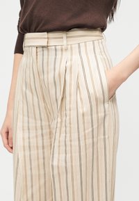 Κρέμ παντελόνι με υψηλή μέση και κάθετες καφέ ρίγες, με αντίστοιχη ζώνη, φορεμένο με καφέ μακρυμάνικο τοπ, το χέρι στην δεξιά τσέπη.