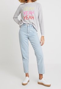 Helle graue Sweatshirt mit buntem "YEAH"-Druck, kombiniert mit blauen High-Waist-Jeans und weißen Sneakers mit beige farbiger Sohle.