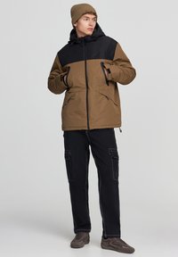 Brązowa i czarna ocieplana kurtka z kapturem, bocznymi kieszeniami i zapięciem na zamek, w połączeniu z czarnymi spodniami cargo i szarymi trampkami.