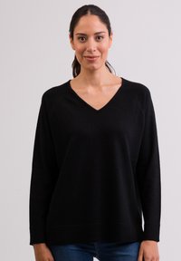 Czarny sweter w serek o miękkiej fakturze, z długimi rękawami raglanowymi i luźnym krojem, noszony na niebieskich dżinsach. Prosty i uniwersalny design.