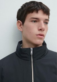 Czarna kurtka na zamek z wysokim kołnierzem, z srebrnym suwakiem. Materiał wydaje się gładki o matowej fakturze. Model ma krótkie włosy.