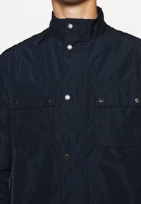 Navy blue, lightweight jacket with a high collar, two chest pockets, and snap button closures. Smooth fabric with a matte finish.
