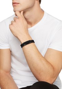 Mustakertainen musta käsikoru, jossa on punottu pinta, litteitä osia ja metallisia yksityiskohtia ranteessa. Yhdistettynä yksinkertaiseen valkoiseen t-paitaan.