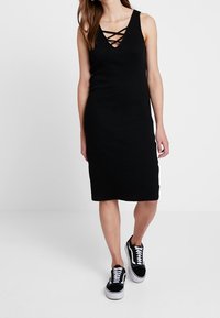 Schwarzes ärmelloses Kleid mit V-Ausschnitt und gekreuzten Trägern, knielang, aus weichem Material, kombiniert mit schwarzen Sneakers.