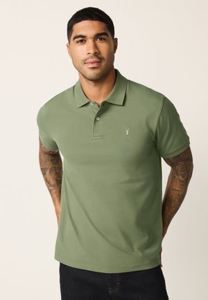 Giovane uomo con capelli corti che indossa una polo verde a maniche corte con tatuaggi visibili su entrambe le braccia.