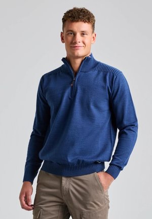 Un homme présente un pull bleu à demi-zip et un pantalon cargo beige sur un fond de studio gris clair.