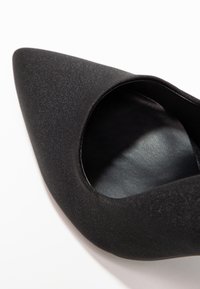 Chaussure à talon haut noire à bout pointu, présentant un extérieur en tissu lisse et un design ajouré. L'intérieur montre une surface noire texturée.