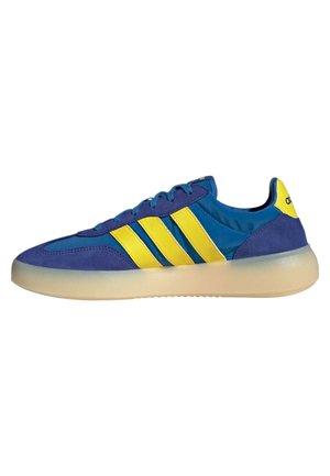 Zapatilla Adidas azul y amarilla con tres rayas amarillas, cordones azules y una suela beige translúcida, vista de lado.