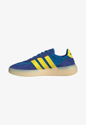 Zapatilla Adidas azul y amarilla con tres rayas amarillas, cordones azules y una suela beige translúcida, vista de lado.