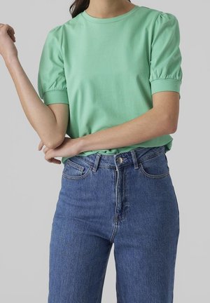 Kvinde iført en mintgrøn bluse med pufærmer og højtaljede blå jeans, med armene krydset om livet.