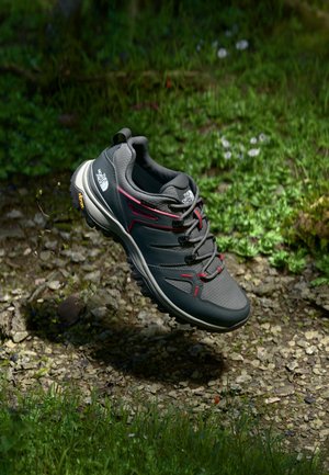 Szare buty trekkingowe z czerwonymi akcentami, wykonane z materiałów tekstylnych i gumowych, z wyściełanym kołnierzem, teksturowaną powierzchnią i podeszwą Vibram, unoszące się nad kamieniami i trawą.