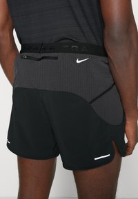 Bermudas de running negras con una cinturilla texturizada en gris, detalles reflectantes y un bolsillo con cremallera. Aberturas de pierna recortadas para mayor movilidad.