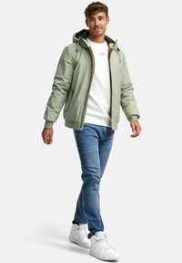 Light green, zip-up jacket with a hood, elastic cuffs, and brown accents. Worn over a white shirt, paired with blue jeans and white sneakers.