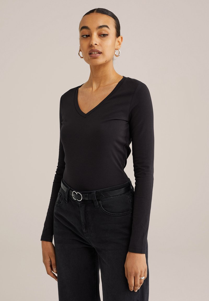 Juodas ilgomis rankovėmis V formos iškirptės marškinėliai, pagaminti iš lygaus audinio, derinami su juodomis aukštu liemeniu džinsais ir diržu, kuriame yra sidabrinė sagtis.