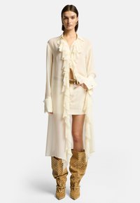 Blusa di crema trasparente con dettagli arricciati, abbinata a una gonna corta di crema e stivali a ginocchio a pois marroni. Accenti in metallo argentato.