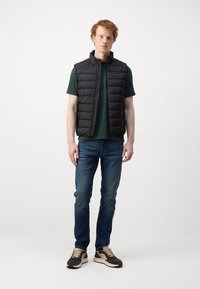 Sort polstret vest med høj krave over en mørkegrøn t-shirt, parret med blå jeans og brune sneakers. Simpelt design og letvægtsmateriale.