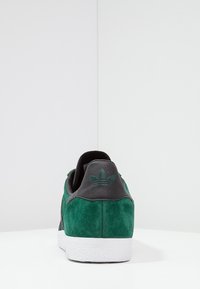 Grön mockasneaker med en svart läderklack och vit gummisula. Inkluderar präglad logga på klacken. Texturerad finish.