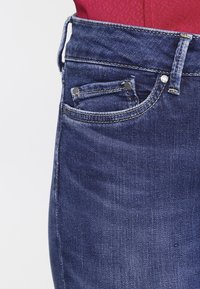 Mörkblå denimjeans med medelhøj midja, sidficka och metalliska detaljer. Tyget har en lätt textur och bleka områden.