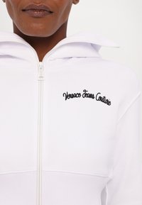 Felpa bianca con zip, caratterizzata dal logo ricamato in nero "Versace Jeans Couture", tessuto morbido, colletto alto e polsini e orlo a coste.