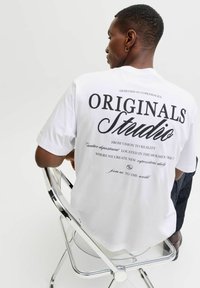 Λευκό βαμβακερό T-shirt με μαύρο κείμενο στο πίσω μέρος, με τη λέξη "ORIGINALS" και "Studio" να εμφανίζονται έντονα. Μοντέλο καθισμένο σε μια χρωμιωμένη καρέκλα.