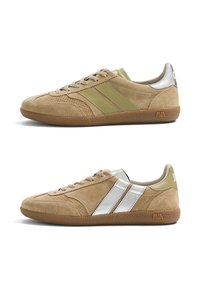 Paire de baskets en daim beige à lacets avec semelles en gomme, l’une avec des rayures vert olive, l’autre avec des rayures argentées et des accents métalliques.