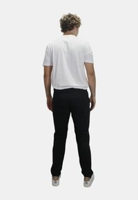 T-shirt bianco a maniche corte, pantaloni neri, girato di spalle. I pantaloni presentano una tasca sul retro; le scarpe sono grigie con accenti marroni. Design semplice e casual.