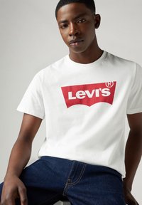 Hvid bomulds t-shirt med et stort rødt Levi's logo centreret på brystet, parret med blå denim jeans. Enkel, klassisk design.