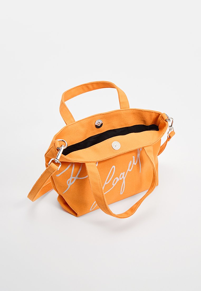 Bolso de lona naranja con dos asas cortas y una correa de hombro desmontable. Presenta herrajes plateados y un detalle de texto blanco en la parte frontal.