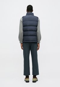 Chaleco de plumas azul marino con cuello alto sobre una camiseta gris de manga larga, combinado con pantalones de pierna ancha verde oscuro y zapatos con acentos marrones.