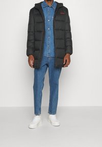 Schwarze Steppjacke mit Kapuze, gesteppter Textur und rotem Logo. Kombiniert mit einem blauen Jeanshemd und lockeren Jeans. Weiße Turnschuhe runden den Look ab.