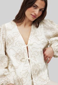 White blouse with beige floral embroidery, deep V-neckline, and tie detail at the center. Elbow-length puff sleeves, soft texture.