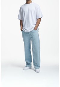 Hombre con jeans sueltos de color azul claro, camiseta blanca de gran tamaño, zapatillas blancas, de pie con las manos en los bolsillos sobre un fondo blanco liso.