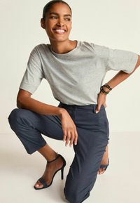 Leende kvinna i knästående med en hand på höften, iklädd grå T-shirt, randiga marinblå byxor, svarta nitararmband och svarta högklackade sandaler.