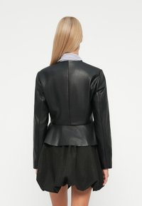 Femme aux longs cheveux blonds portant une veste en cuir noir ajustée sur une jupe noire avec un ourlet bouffant, vue de dos.
