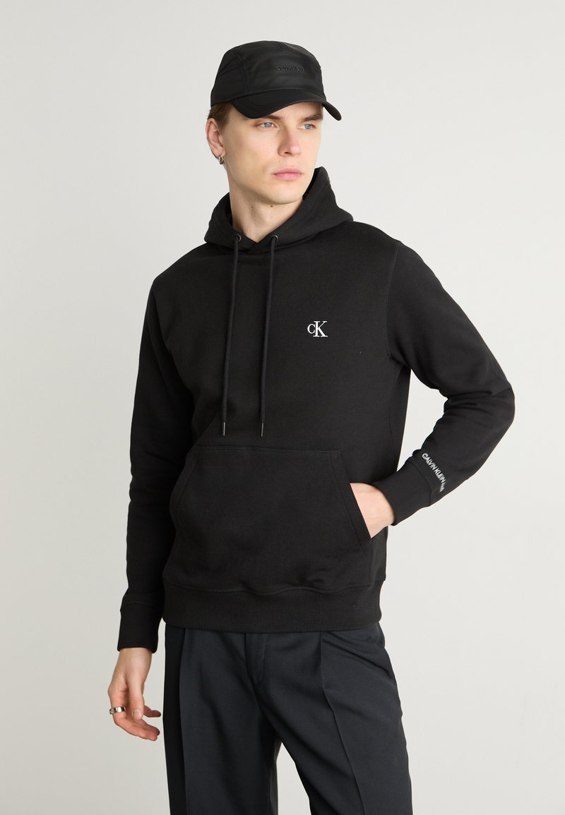 Hoodie en coton noir avec une poche frontale, capuche à cordon de serrage et logo brodé. Associe avec un pantalon noir et une casquette noire.