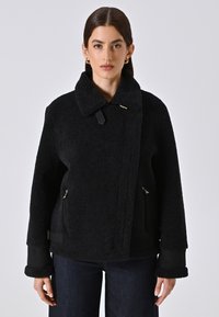 Giacca nera dalla texture unica con colletto, tasche con zip e polsini a contrasto. Presenta un accento dorato e una chiusura frontale asimmetrica.
