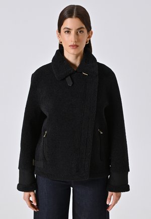 Schwarze, strukturierte Jacke mit Kragen, Reißverschlusstaschen und kontrastierenden Manschetten. Besondere Merkmale sind ein goldener Akzent und ein asymmetrischer Frontverschluss.