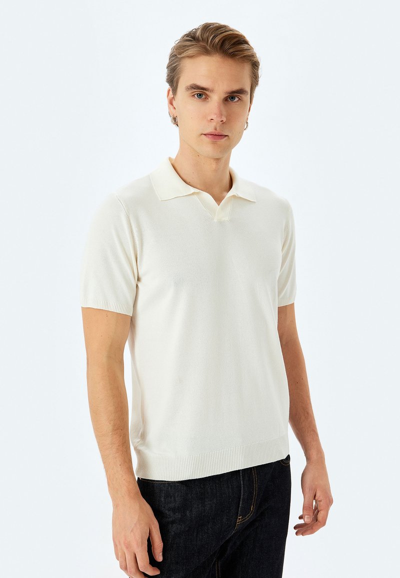 Kortærmet cremefarvet polo med ribbede krave og kant, der har en glat tekstur og tætsiddende design, parret med mørke denimjeans.