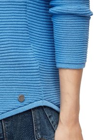 Pull en tricot bleu clair avec une texture côtelée, présentant un ourlet arrondi, des manches trois-quarts et un petit détail de logo métallique.